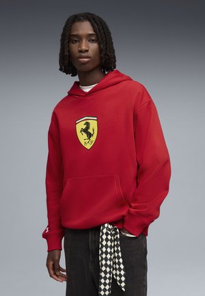 Rotes Kapuzen-Sweatshirt mit großem gelbem Ferrari-Logo. Verfügt über eine Fronttasche und gerippte Bündchen, kombiniert mit dunkler Jeans und einem Schal mit Schachbrettmuster.