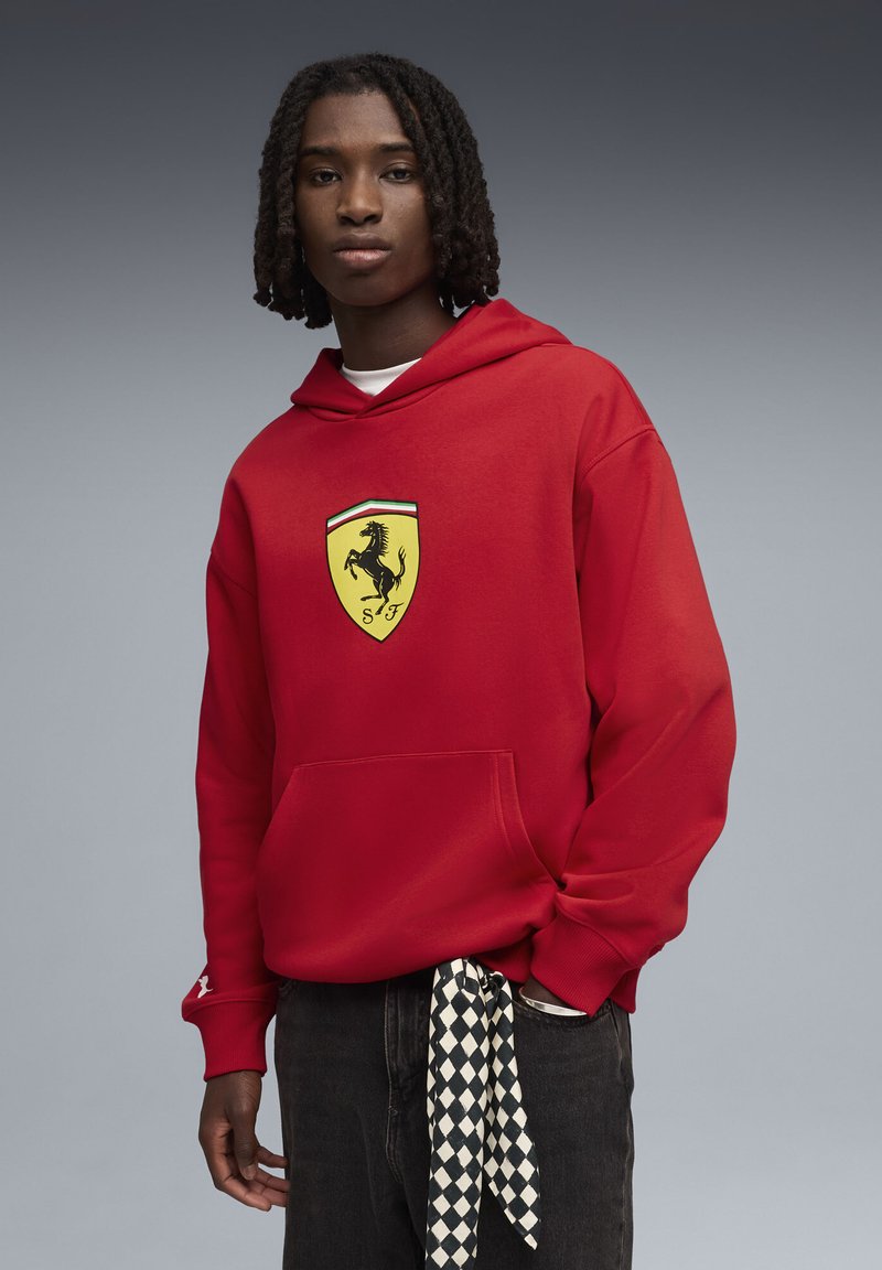 Rød hættetrøje med stort gult Ferrari-logo. Har en forlommer og ribbede manchetter, kombineret med mørke jeans og et halstørklæde med ternet mønster.