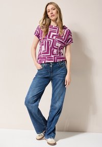 Kortærmet lila top med et geometrisk mønster, parret med løstsiddende blå jeans og beige sneakers. Glat stof, afslappet design.
