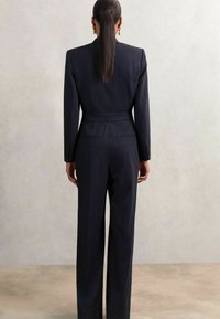 Donna con capelli lisci raccolti in una coda bassa, che indossa un completo pantalone nero a righe sottili, in piedi di fronte a una parete texturizzata grigio chiaro.