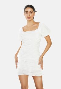 Mujer de cabello oscuro con un mini vestido blanco fruncido y mangas cortas abullonadas, de pie frente a un fondo claro y liso.