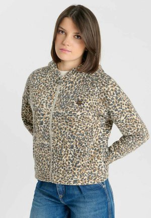Giovane donna con capelli castani lisci che indossa una giacca beige e grigia con stampa leopardata con cerniera e jeans blu, in piedi con le mani dietro la schiena.