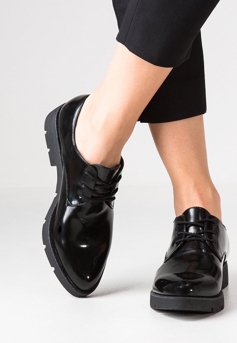 Chaussures en cuir noir brillant à lacets portées avec un pantalon noir raccourci, jambes croisées sur un fond clair uni.