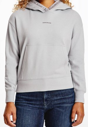 Jasnoszara bluza z kapturem z przednią kieszenią i logo "Calvin Klein Jeans" na piersi oraz kapturze, noszona z ciemnoniebieskimi dżinsami.