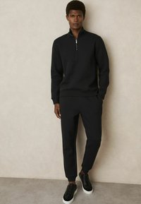 Pulover din tricot negru cu guler cu fermoar pe jumătate, asortat cu pantaloni joggeri tapered și adidași negri cu tălpi albe.