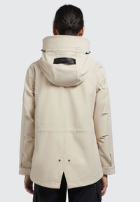 Beige vattentät jacka med hög krage, justerbar huva och svart logotag. Har en ventil på baksidan och dragsko för passformsjustering.
