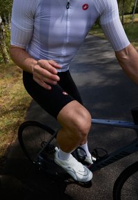 Biała koszulka rowerowa z krótkim rękawem, czarne spodenki rowerowe i białe buty. Rowerzysta znajduje się na szarym rowerze na utwardzonej ścieżce.