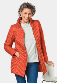 Gequilte, oranje jas met een hoge kraag, een rits aan de voorkant en twee zijzakken. Gekocht over een lichtgrijze top en gecombineerd met blauwe jeans.