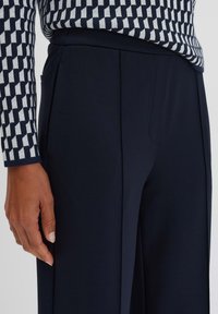 Pantaloni su misura blu navy con una texture liscia, caratterizzati da pieghe frontali e tasche laterali. Un maglione a fantasia con design geometrici è visibile.
