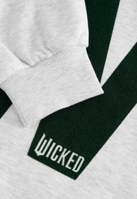 Szary materiał bluzy z prążkowanymi mankietami, z ciemnozielonymi akcentami i logo "WICKED" umieszczonym blisko mankietu. Miękka tekstura z gładkim wykończeniem.