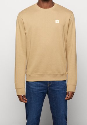 Sudadera de cuello redondo beige con mangas largas, puños acanalados y un pequeño parche con logotipo en el pecho, combinado con jeans azul oscuro.