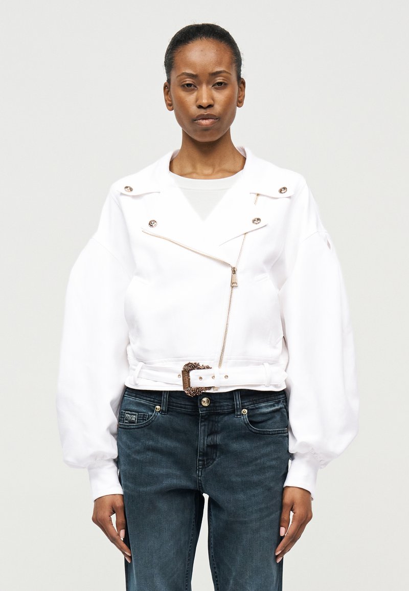 Γυναίκα με μαλλιά δεμένα πίσω φοράει λευκό υπερμεγέθες μπουφάν με φερμουάρ και ζώνη, συνδυασμένο με σκούρο μπλε τζιν, όρθια μπροστά σε απλό φόντο.