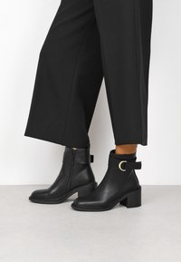 Botines de cuero negro con cierre de cremallera, tacón cuadrado y una correa lateral con un acento redondo dorado. Textura lisa y diseño estilizado.
