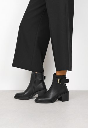 Botines de cuero negro con cierre de cremallera, tacón cuadrado y una correa lateral con un acento redondo dorado. Textura lisa y diseño estilizado.
