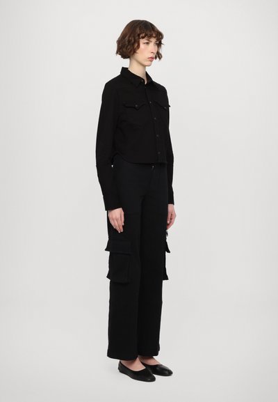 Camicia nera croppata con bottoni e due tasche sul petto, abbinata a pantaloni cargo neri a gamba larga con tasche laterali. Scarpe basse nere.