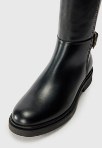 Botte noire en cuir jusqu'au genou avec une texture lisse, bout rond, fermeture éclair sur le côté et un petit talon empilé. Accentuée d'une boucle métallique.