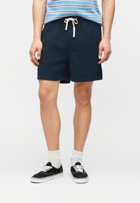 Άνδρας που φοράει μαύρα σορτς με κορδόνι, άσπρες κάλτσες, μαύρα αθλητικά παπούτσια και μπλε ριγέ πουκάμισο, στέκεται μπροστά σε απλό φόντο.