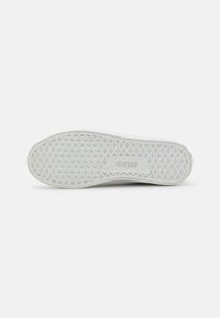 Suela de zapatilla de goma blanca con patrón de diamante texturizado y surcos. Presenta el logo "GUESS" en el talón. Forma lisa y plana.