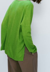 Personne portant un haut à manches longues vert vif et un pantalon marron, les mains dans les poches, debout devant un fond clair uni.