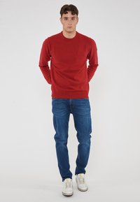 Giovane in piedi con le mani dietro la schiena, indossando un maglione rosso, jeans blu e sneaker bianche, su uno sfondo chiaro e semplice.