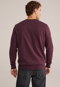 Uomo con capelli corti e ricci, che indossa un semplice felpa bordeaux e jeans grigi scuri, in piedi con le spalle rivolte verso un sfondo chiaro.
