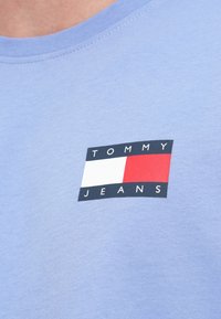 T-shirt azul claro com decote redondo. Apresenta um logo retangular preto com um gráfico em branco e vermelho e o texto "TOMMY JEANS."