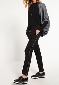 Persona che indossa una camicia nera con bottoni, pantaloni neri con strisce bianche ai lati, scarpe platform nere e un cardigan grigio aperto, in piedi con le mani in tasca.