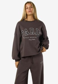 Giovane donna con lunghi capelli biondi che indossa una felpa over-size scura e pantaloni abbinati con il testo "PARIS" su entrambi i capi.