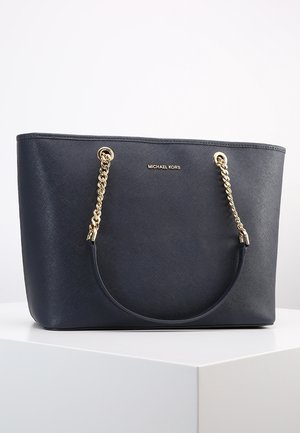 Sort Michael Kors tote taske med gyldne kædehåndtag, der hviler på en hvid overflade mod en ensfarvet baggrund.