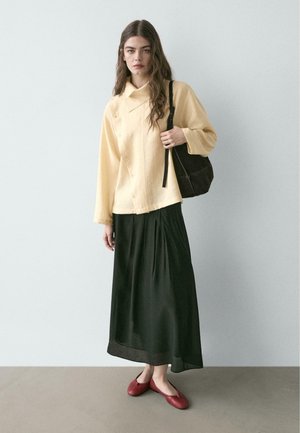 Femme aux longs cheveux bruns portant un chemisier boutonné jaune pâle, une jupe plissée noire, des ballerines rouges, et portant un sac à bandoulière noir.