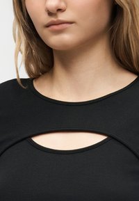 Γυναίκα που φοράει μαύρη μπλούζα με στρογγυλεμένη κοπή κάτω από το λαιμό, αφήνοντας εμφανές ένα τμήμα δέρματος στο πάνω μέρος του στήθους.