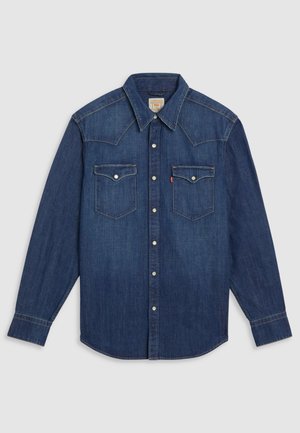 Donkerblauw denim shirt met lange mouwen, puntige kraag, witte drukknoopjes en twee borstzakken. Voorzien van gestikte schouderdetails.