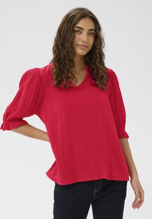 Młoda kobieta o kręconych brązowych włosach, ubrana w jasnoczerwoną, fakturowaną bluzkę z bufiastymi rękawami i ciemne jeansy, stoi na tle jednolitego tła.