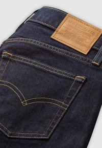 Calças de ganga escuras com uma etiqueta de couro castanha. Costuras amarelas contrastantes e design clássico do bolso traseiro com um detalhe de costura curva.