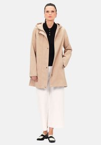 Cappotto impermeabile beige con cappuccio, zip frontale e chiusure a scatto, abbinato a una maglia nera e pantaloni bianchi a gamba larga.