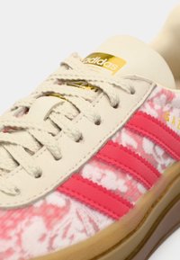 Adidas Gazelle sneaker featuring cream upper, pink suede stripes, and a textured pink and white pattern. Gold logo accent on tongue.