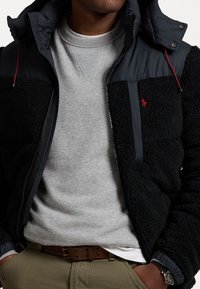 Man som bär en svart fleecejacka över en grå sweatshirt, olivgröna byxor med brunt bälte, händer i fickorna och armbandsur på vänster handled.