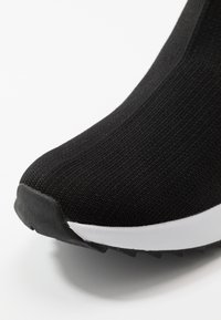 Musta urheilukenkä, jossa on teksturoitu neulottu päällinen, valkoinen kumipohja ja musta pito-ulkopohja. Lähikuva kärkiosasta.