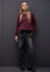 Sudadera marrón con mangas largas, combinada con jeans negros de pierna recta. El modelo está de pie contra una pared oscura, llevando botas negras hasta el tobillo.
