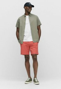 Kortärmad, olivgrön skjorta med knappar över en vit t-shirt, i kombination med korallfärgade shorts och olivgröna sneakers, prydd med en marinblå keps.