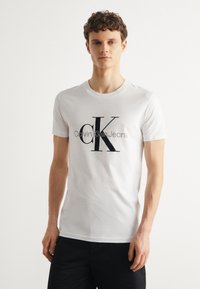 Balta kokvilnas T-kreklā ir liels melns "CK" logo un teksts "Calvin Klein Jeans". Ar īsām piedurknēm un apaļu kakla izgriezumu.