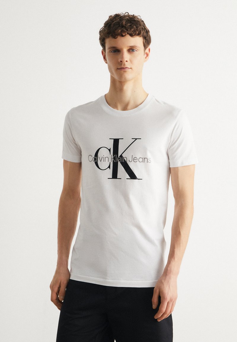 Balta kokvilnas T-kreklā ir liels melns "CK" logo un teksts "Calvin Klein Jeans". Ar īsām piedurknēm un apaļu kakla izgriezumu.