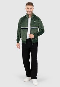 Giacca a track in velluto verde con colletto alto, strisce orizzontali bianche e blu navy, e logo. Abbinata a pantaloni neri e sneaker bianche.