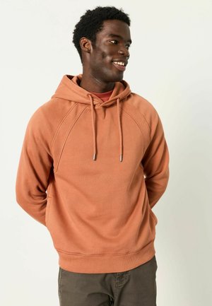 Lächelnder Mann, der seitlich schaut, trägt einen rostfarbenen Hoodie und dunkle Hosen, steht mit den Händen hinter dem Rücken vor einem einfarbigen Hintergrund.