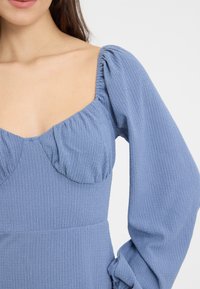 Blå teksturert kjole med firkantet utringning, puffede lange ermer og rynkede detaljer ved brystet. Stoffet ser lett og litt krøllete ut.