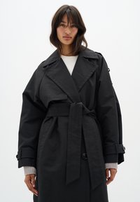Un manteau trench noir avec une taille ceinturée, un grand col et des poignets boutonnés, en tissu lisse. Superposé sur un haut léger.