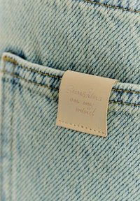 Džínovina v světle modrých tónech, s koženou cedulkou přišitou na kapse, na které je vyražen nápis "Sluneční paprsek v mé mysli".