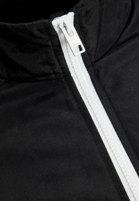 Giacca nera con colletto alto, chiusura a zip bianca e texture della stoffa liscia. Il design è minimalista, senza pattern visibili.