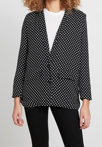 Czarna marynarka w białe grochy, z długimi rękawami, klapami z wycięciami, dwoma przednimi kieszeniami i pojedynczym zapięciem na guzik. Gładka faktura tkaniny.