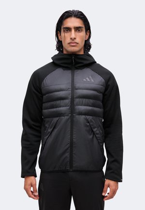 Mann mit schwarzen schulterlangen Haaren, der eine schwarze Zip-Up-Jacke mit gepolstertem Vorderteil und Adidas-Logo auf Brust und Hose trägt.